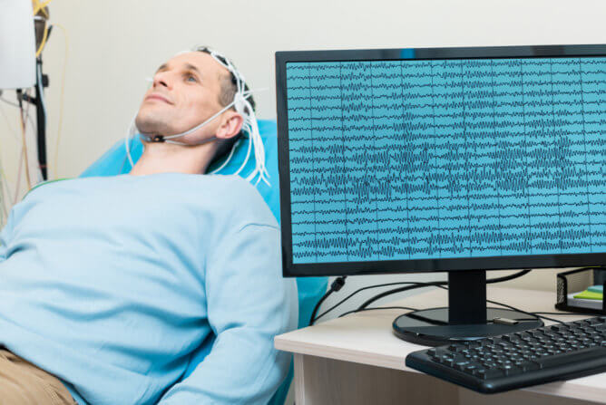 A man is lying down in a clinic while a computer shows his brain activity from an EEG test.