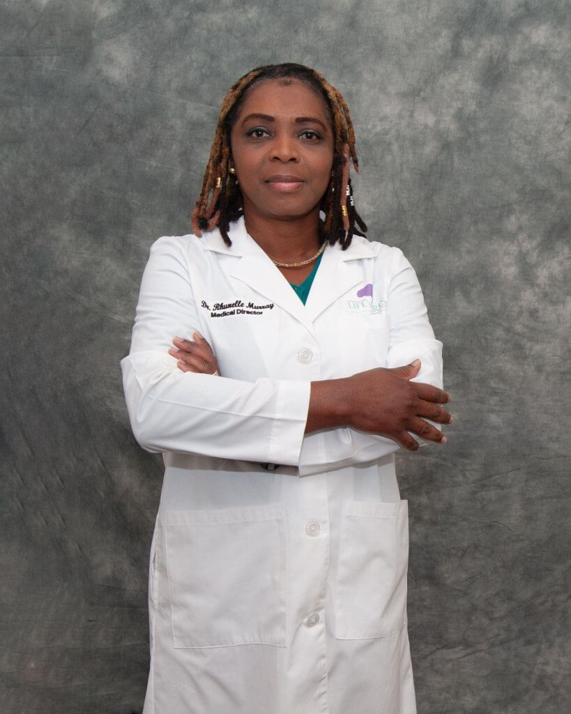 A person wearing a white lab coat with arms crossed, standing against a gray textured backdrop.