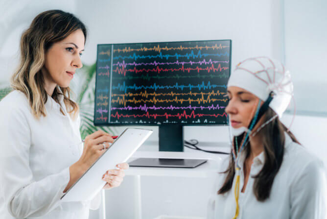 A woman measures a woman's brain activity using an electroencephalogram in a clinical setting.